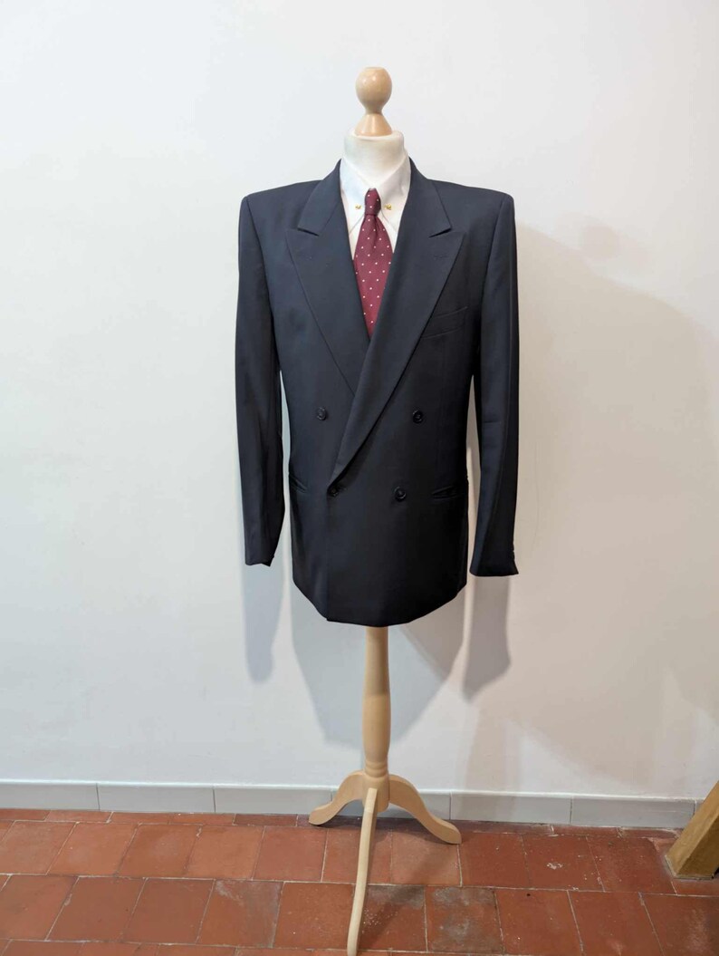 May include: A dark navy double-breasted blazer, white collared shirt, and burgundy patterned tie on a wooden mannequin. The blazer features a classic design with notched lapels and a double-button closure. The suit is a formal wear item.