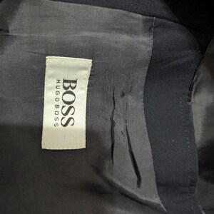 May include: Close-up of the interior lining of a dark navy blue blazer. A white tag with the brand name "BOSS HUGO BOSS" is sewn into the lining. The lining is a smooth, dark fabric.