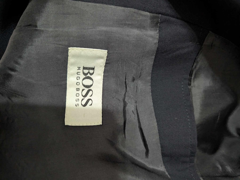 May include: Close-up of the interior lining of a dark navy blue blazer. A white tag with the brand name "BOSS HUGO BOSS" is sewn into the lining. The lining is a smooth, dark fabric.