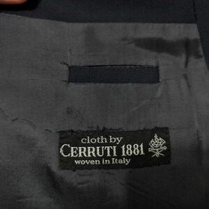 May include: Close-up of the interior lining of a dark navy blue suit jacket. The lining is a dark grey fabric with a small pocket. A black label reads "cloth by CERRUTI 1881 woven in Italy."