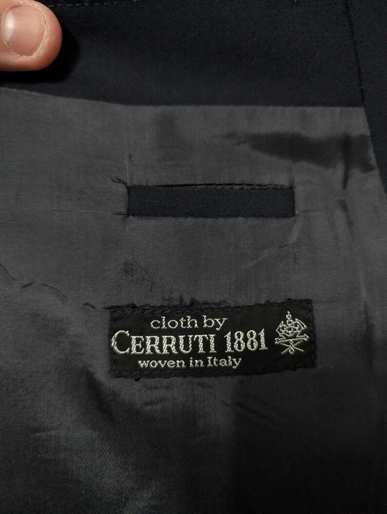 May include: Close-up of the interior lining of a dark navy blue suit jacket. The lining is a dark grey fabric with a small pocket. A black label reads "cloth by CERRUTI 1881 woven in Italy."