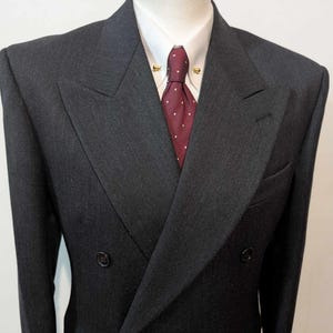 Puede incluir: Chaqueta de traje cruzada gris carb&oacute;n con camisa blanca de cuello y corbata burdeos con lunares blancos. La chaqueta tiene dos filas de botones y solapa con muescas.