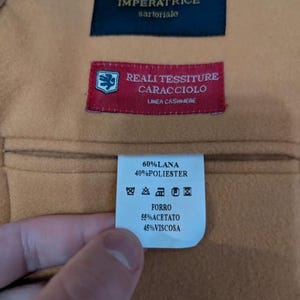 Puede incluir: Primer plano de una tela beige con tres etiquetas cosidas. La etiqueta superior es azul marino con texto dorado, la etiqueta central es roja con texto blanco y la etiqueta inferior es blanca con texto negro. Las etiquetas proporcionan informaci&oacute;n sobre la composici&oacute;n de la tela.