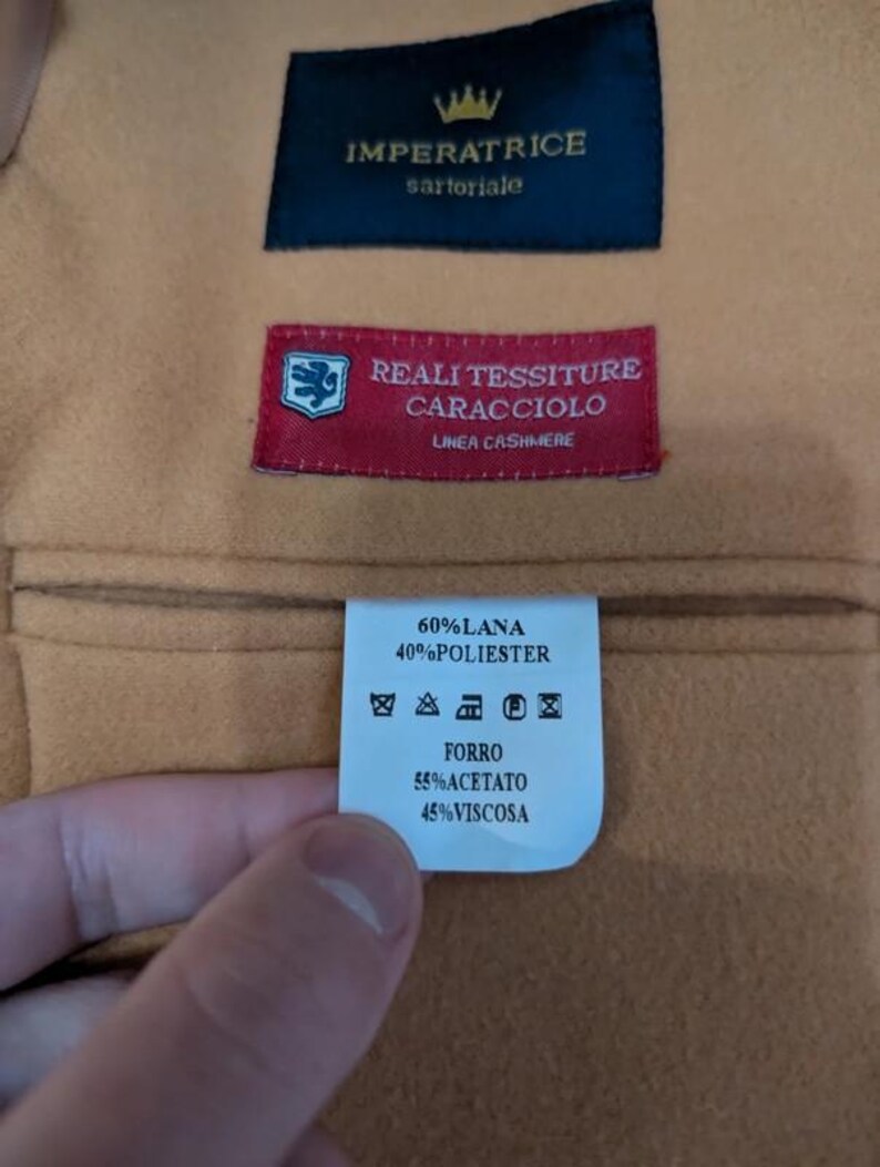 Puede incluir: Primer plano de una tela beige con tres etiquetas cosidas. La etiqueta superior es azul marino con texto dorado, la etiqueta central es roja con texto blanco y la etiqueta inferior es blanca con texto negro. Las etiquetas proporcionan informaci&oacute;n sobre la composici&oacute;n de la tela.