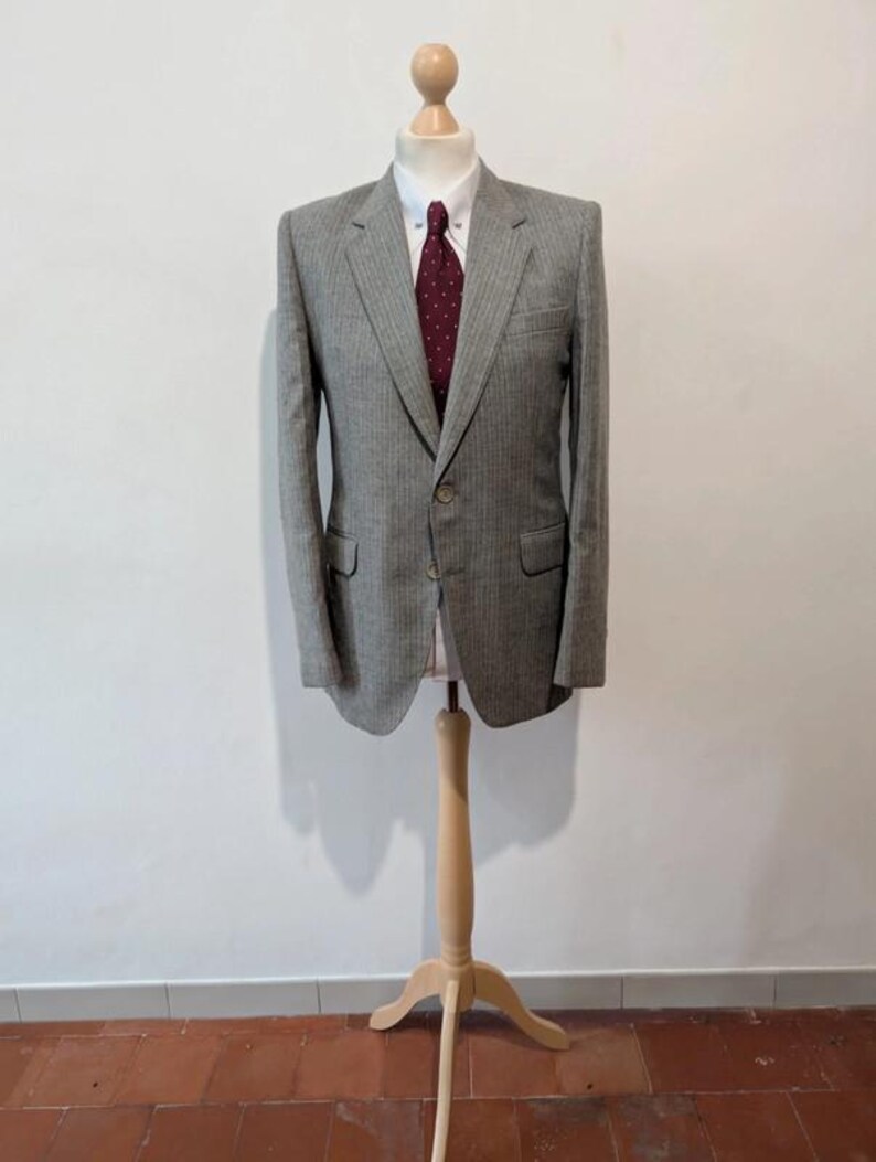 May include: A gray pinstripe blazer with a white collared shirt and a burgundy tie. The blazer has two front pockets and a single button closure. The outfit is displayed on a wooden mannequin.