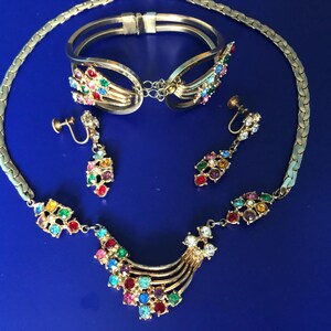 Puede incluir: Un conjunto de joyas vintage con un collar, una pulsera y pendientes dorados. El conjunto está adornado con piedras preciosas redondas y coloridas. El collar y la pulsera tienen una cadena plateada. Los pendientes son de clip.