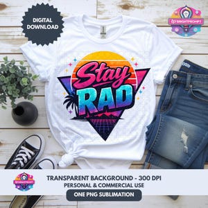 May include: White t-shirt with a colorful "Stay Rad" graphic design. The design features a retro sunset, palm tree, and geometric shapes. The shirt is paired with blue jeans and black sneakers, suggesting a casual, everyday style.