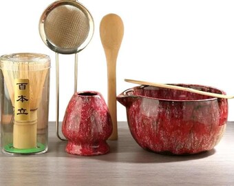 May include: A matcha tea set with a bamboo whisk in a clear container, a metal sieve, a wooden spoon, a red ceramic bowl, a red ceramic holder, and a bamboo scoop. The container has Japanese text.