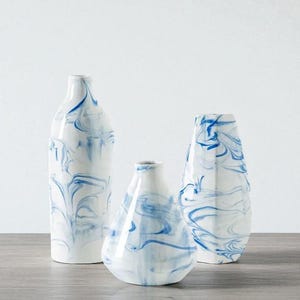 May include: Three white ceramic vases with a blue swirled pattern. The vases are different shapes and sizes, with a tall, slender bottle shape, a rounded teardrop shape, and a faceted, slightly flared shape. The vases are displayed on a grey surface.