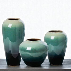 Può includere: Tre vasi in ceramica di varie dimensioni e forme. I vasi presentano una combinazione di colori sfumati, che vanno da un verde-blu scuro alla base a un verde-blu più chiaro e crema nella parte superiore. I bordi sono marrone scuro.