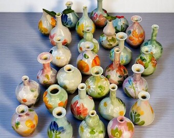 Pode incluir: Uma coleção de pequenos vasos de cerâmica pintados à mão em várias formas e tamanhos. Os vasos apresentam desenhos florais e abstratos coloridos em tons de laranja, verde, azul e rosa. Alguns têm formato bulboso, outros são mais alongados.