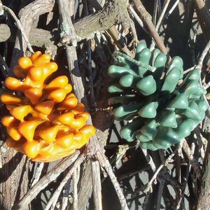 May include: Two pine cone shaped candles, one is orange and the other is green. The candles are sitting on a bed of twigs.