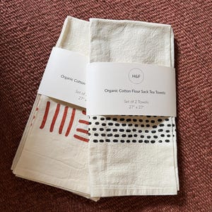 May include: Two organic cotton flour sack tea towels, each 27" x 27", are stacked and wrapped with a paper band. One towel has a red dash design, the other has a black dotted pattern. The paper band reads "Organic Cotton Flour Sack Tea Towels".