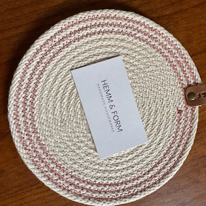 May include: A round, woven coaster with a cream-colored base and red stitching. A white card with the text "HEMM & FORM HANDMADE HOUSEWARES" rests on the coaster. A small leather tag with "H&F" is attached.