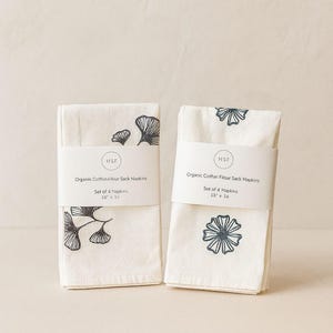 May include: Two sets of white organic cotton flour sack napkins, each wrapped with a paper band. The napkins feature a dark blue floral design. Each set contains four napkins, measuring 18 x 16 inches.
