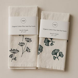 May include: Two off-white organic cotton flour sack tea towels, each 27 x 27 inches, with black botanical designs. One towel features a floral pattern, the other has a leaf design. Each towel is labeled with the text "H&F" and "Organic Cotton Flour Sack Tea Towels."