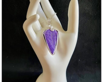 May include: A silver pendant necklace featuring a polished, shield-shaped purple gemstone. The gemstone is set in a silver wire frame and hangs from a delicate silver chain. The pendant is displayed on a white hand-shaped jewelry display.