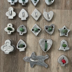 Può includere: Una collezione di 16 tagliapasta vintage in metallo di varie forme, tra cui cuori, stelle, campane, omini di pan di zenzero e semi di carte da gioco. I tagliapasta hanno manici in legno verde e rosso.