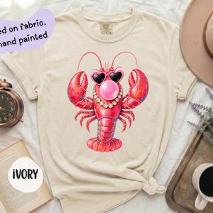 May include: An ivory t-shirt featuring a graphic of a red lobster wearing heart-shaped sunglasses, a pearl necklace, and blowing a pink bubble. The text on the shirt reads Printed on fabric. Not hand painted. The shirt is ivory in color.