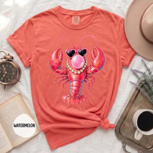 May include: A coral-colored t-shirt featuring a graphic of a red lobster wearing heart-shaped sunglasses, a pearl necklace, and blowing a pink bubblegum bubble. The shirt has short sleeves, a crew neckline, and a folded hem at the sleeve ends.