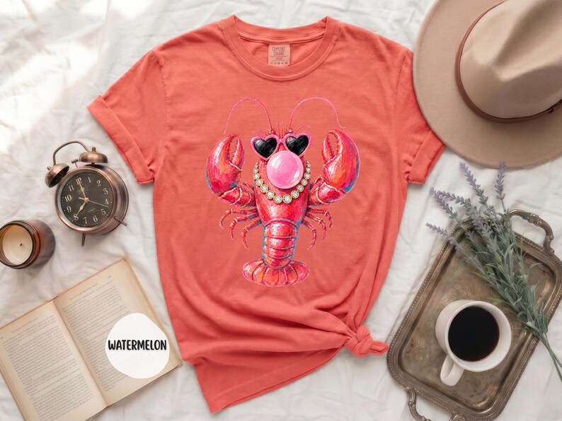 May include: A coral-colored t-shirt featuring a graphic of a red lobster wearing heart-shaped sunglasses, a pearl necklace, and blowing a pink bubblegum bubble. The shirt has short sleeves, a crew neckline, and a folded hem at the sleeve ends.