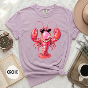 May include: A light purple short-sleeve t-shirt featuring a graphic of a red lobster wearing heart-shaped sunglasses and a pearl necklace while blowing a pink bubble. The shirt has a crew neckline and a label printed on the inside collar area.