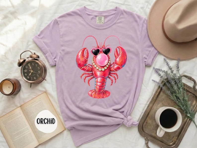 May include: A light purple short-sleeve t-shirt featuring a graphic of a red lobster wearing heart-shaped sunglasses and a pearl necklace while blowing a pink bubble. The shirt has a crew neckline and a label printed on the inside collar area.