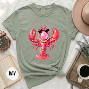 May include: A sage green t-shirt featuring a central graphic of a bright red lobster wearing black heart-shaped sunglasses, a pearl necklace, and blowing a pink bubblegum bubble. The shirt has short sleeves and a crew neckline, presented in a flat lay.