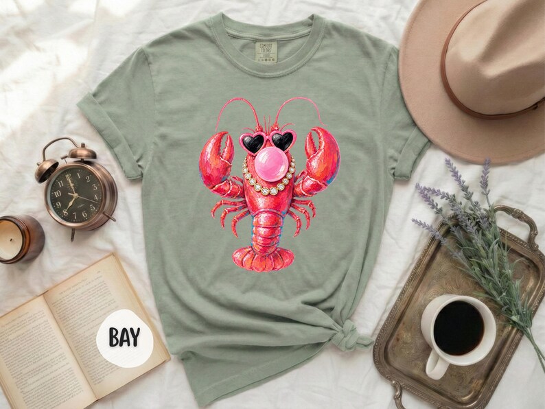 May include: A sage green t-shirt featuring a central graphic of a bright red lobster wearing black heart-shaped sunglasses, a pearl necklace, and blowing a pink bubblegum bubble. The shirt has short sleeves and a crew neckline, presented in a flat lay.