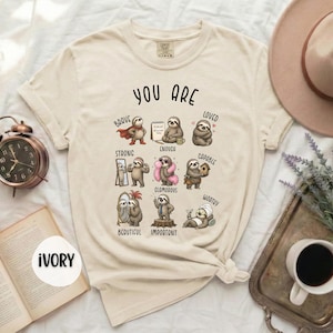 May include: An ivory t-shirt featuring a graphic print with the text "YOU ARE" above nine illustrations of sloths. Each sloth is labelled with words including "BRAVE," "LOVED," "STRONG," "ENOUGH," "CAPABLE," "BEAUTIFUL," "IMPORTANT," and "WORTHY."