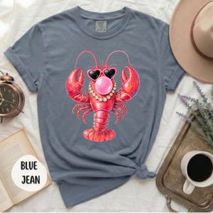 May include: A blue jean colored t-shirt featuring a graphic of a red lobster wearing heart-shaped sunglasses, a pearl necklace, and blowing a pink bubble. The shirt has short sleeves and a crew neckline, with the text BLUE JEAN printed on a circular tag.