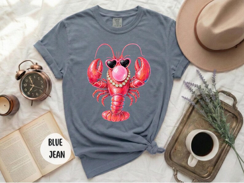 May include: A blue jean colored t-shirt featuring a graphic of a red lobster wearing heart-shaped sunglasses, a pearl necklace, and blowing a pink bubble. The shirt has short sleeves and a crew neckline, with the text BLUE JEAN printed on a circular tag.