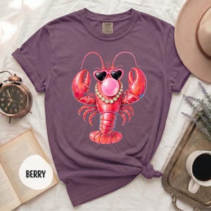 May include: A berry-colored t-shirt featuring a graphic of a red lobster wearing heart-shaped sunglasses, a pearl necklace, and blowing a pink bubble. The shirt has short sleeves and a crew neckline, with the word BERRY printed on a white circle.