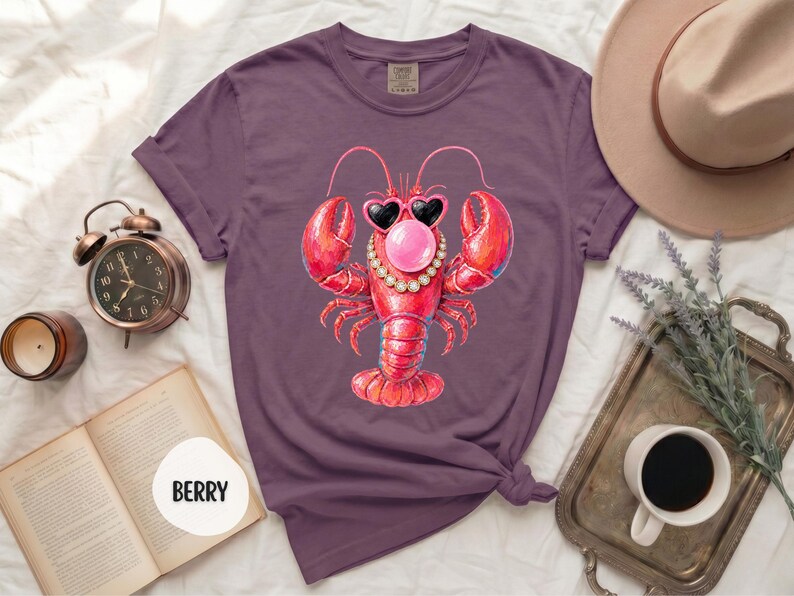 May include: A berry-colored t-shirt featuring a graphic of a red lobster wearing heart-shaped sunglasses, a pearl necklace, and blowing a pink bubble. The shirt has short sleeves and a crew neckline, with the word BERRY printed on a white circle.