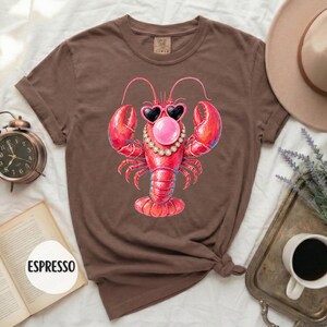 May include: A brown short-sleeve t-shirt featuring a graphic of a red lobster wearing heart-shaped sunglasses, a pearl necklace, and blowing a pink bubble. The shirt has a crew neckline and a label at the inner collar. The text ESPRESSO is visible.