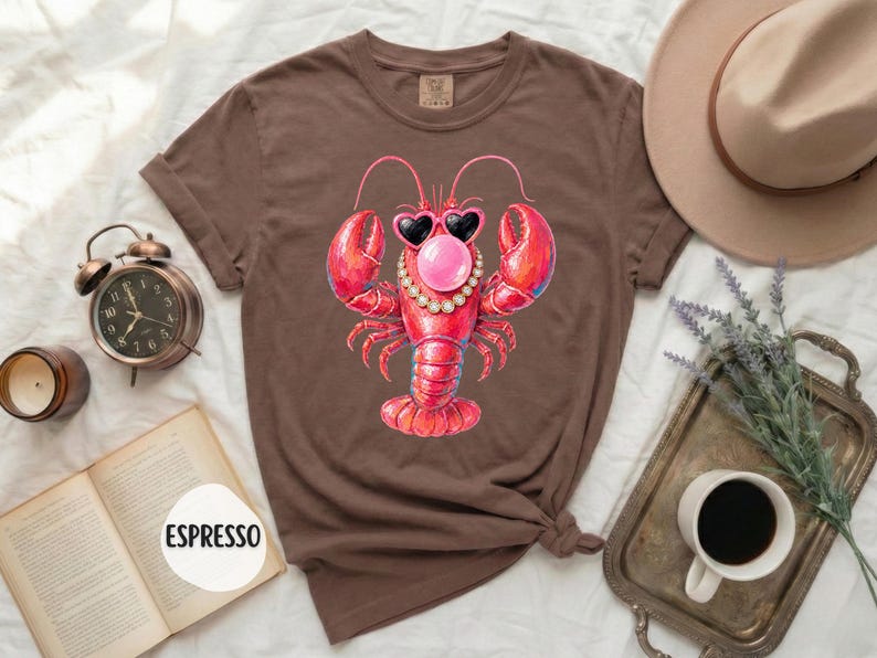 May include: A brown short-sleeve t-shirt featuring a graphic of a red lobster wearing heart-shaped sunglasses, a pearl necklace, and blowing a pink bubble. The shirt has a crew neckline and a label at the inner collar. The text ESPRESSO is visible.