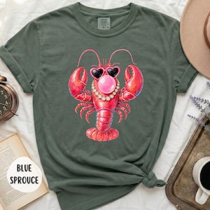 May include: A dark olive green t-shirt featuring a graphic of a red lobster wearing heart-shaped sunglasses, a pearl necklace, and blowing a pink bubble gum bubble. The shirt has short sleeves and a crew neckline, with the text BLUE SPROUCE visible.