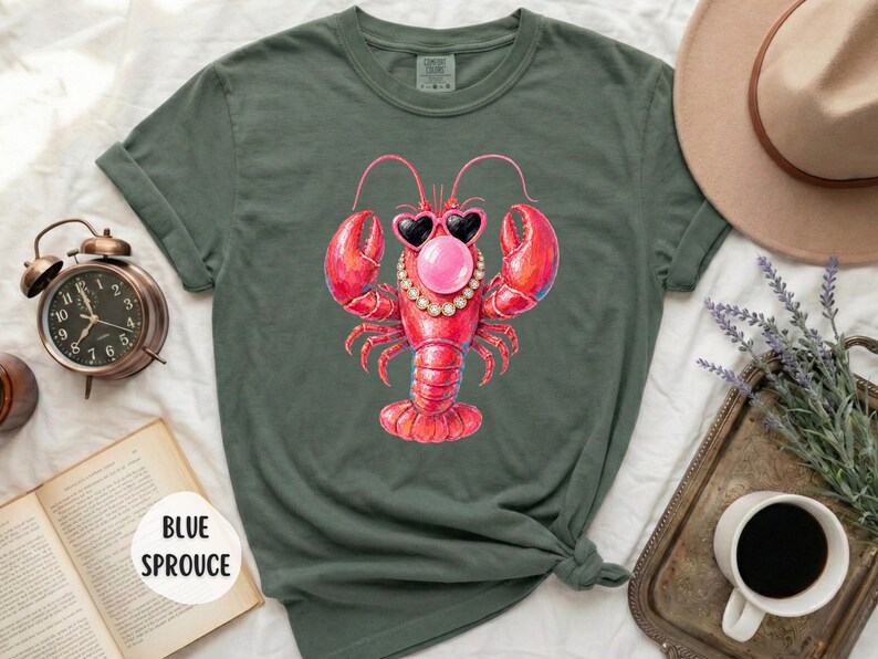 May include: A dark olive green t-shirt featuring a graphic of a red lobster wearing heart-shaped sunglasses, a pearl necklace, and blowing a pink bubble gum bubble. The shirt has short sleeves and a crew neckline, with the text BLUE SPROUCE visible.