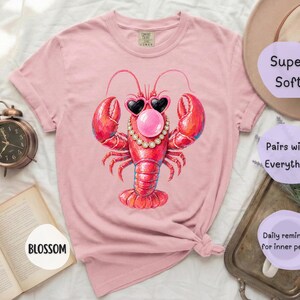 May include: A light pink short-sleeve t-shirt featuring a graphic of a red lobster wearing heart-shaped sunglasses, a pearl necklace, and blowing a pink bubble. The shirt has a crew neckline and a relaxed fit with a visible brand tag at the collar.