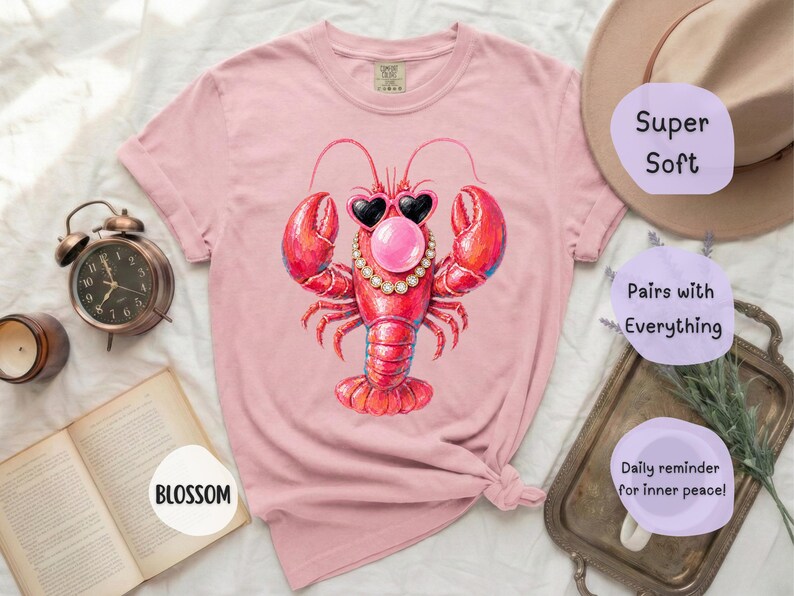 May include: A light pink short-sleeve t-shirt featuring a graphic of a red lobster wearing heart-shaped sunglasses, a pearl necklace, and blowing a pink bubble. The shirt has a crew neckline and a relaxed fit with a visible brand tag at the collar.