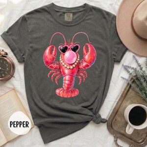 May include: A charcoal gray short-sleeve t-shirt featuring a graphic of a red lobster wearing heart-shaped sunglasses and a beaded necklace while blowing a pink bubble. The shirt has a crew neckline and rolled sleeves, with the word PEPPER visible.