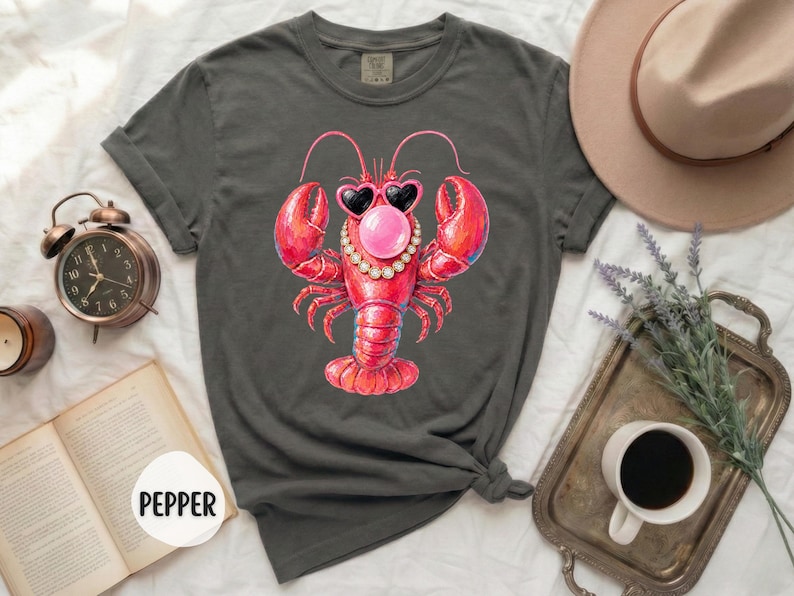 May include: A charcoal gray short-sleeve t-shirt featuring a graphic of a red lobster wearing heart-shaped sunglasses and a beaded necklace while blowing a pink bubble. The shirt has a crew neckline and rolled sleeves, with the word PEPPER visible.
