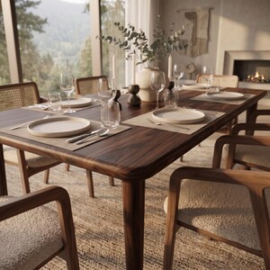 May include: A dark wood dining table set with cream-colored ceramic plates, clear glass tumblers, wine glasses, and silver cutlery on rectangular placemats. A central ceramic vase holds eucalyptus branches, flanked by two tall white taper candles.