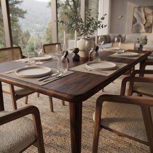 May include: A dark wood dining table set with light-colored plates, clear glass tumblers, wine glasses, and silver cutlery on rectangular placemats. The table is surrounded by wooden chairs with woven cane backrests and textured beige seat cushions.