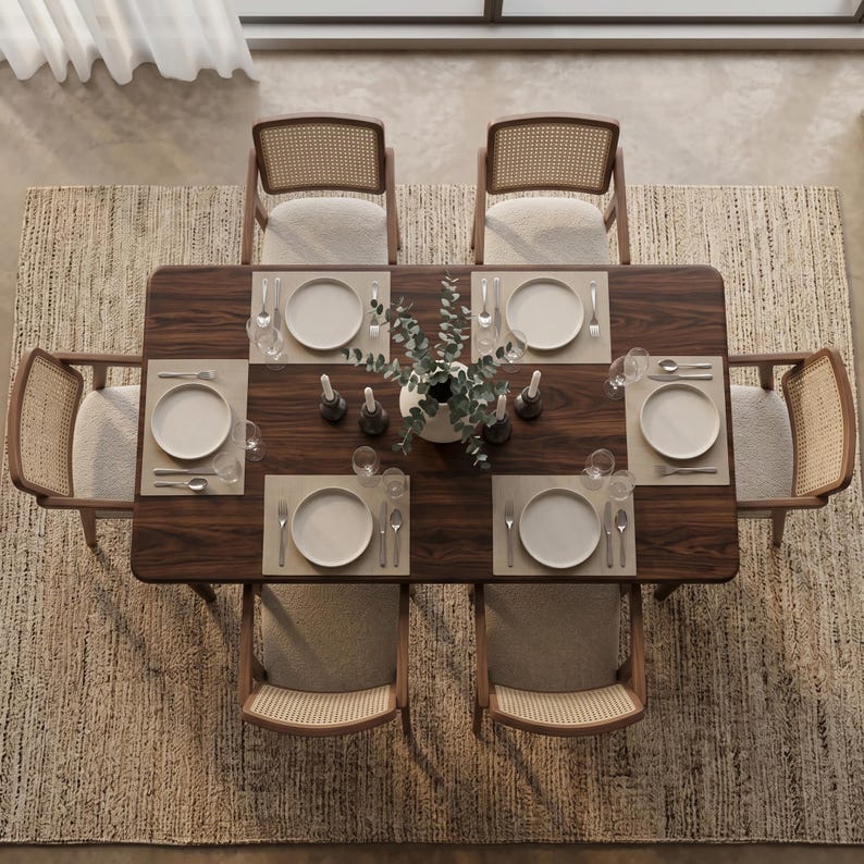 May include: A rectangular dark wood dining table with six chairs featuring light-colored upholstered seats and woven cane backrests. The table is set with six white plates, silverware, glassware, and a central vase with green foliage.
