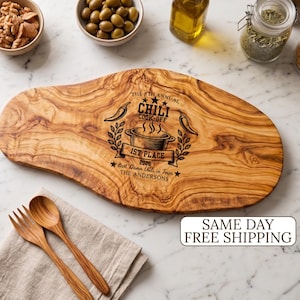 May include: An oval-shaped wooden serving board with a natural grain pattern, engraved with the text THE 5TH ANNUAL CHILI COOK-OFF 1ST PLACE 2026 BEST DAMN CHILI IN TOWN THE ANDERSONS, featuring chili peppers and a pot. Includes a wooden fork and spoon.