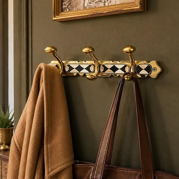 May include: A wall-mounted brass coat rack featuring three curved hooks and a decorative horizontal plate with a repeating black and white diamond pattern. A tan coat and a brown leather bag are hanging from the hooks on the metallic fixture.