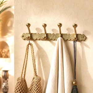 Könnte beinhalten: Eine wandmontierte Garderobenleiste aus Messing mit vier nach oben gebogenen Haken, die an einer horizontalen Metallplatte mit geprägtem Blumenmuster befestigt sind. Eine beige geflochtene Tasche, ein weißes Tuch und eine dunkelgrüne Quaste hängen daran.