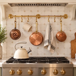 Puede incluir: Una barra de cocina de latón montada en una pared de azulejos con ganchos en S que sostienen dos sartenes de cobre, una taza blanca acanalada y un paño de cocina a rayas blancas y grises. Un hervidor de color crema sobre una estufa de gas.