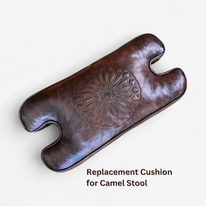 May include: A dark brown leather replacement cushion for a camel stool featuring a central embossed geometric star pattern and decorative floral motifs on the ends. The text Replacement Cushion for Camel Stool is printed in brown below the item.